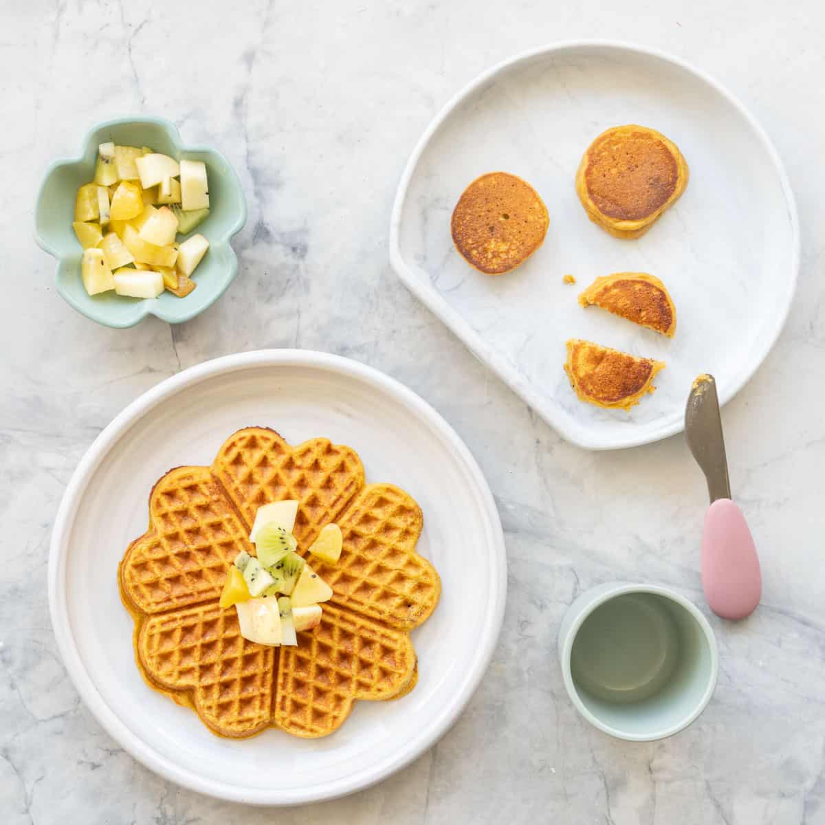 A pumpkin waffle, 3 baby pancakes and a bowl of fruit on a bench top.