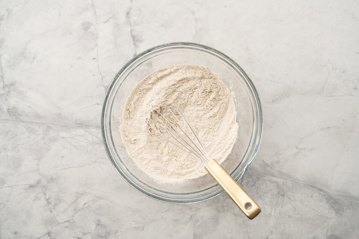 A glass mixing bowl of dry ingredients with a brass handles whisk resting on the side.