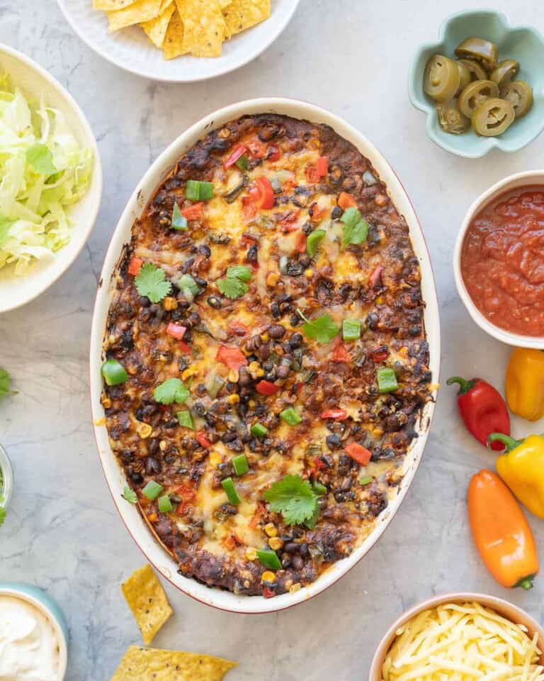 A casserole topped with melted cheese, diced capsicum, corn and beans on a bench top scattered with tortilla chips and coriander leaves.