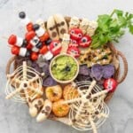 Halloween snack board on benchtop with many different halloween themed food. Cheese Monster, Mummy Cookies, Ants on a log, Spiderweb Pretzels, Fruit skewers, Banana Ghosts, Mandarin pumpkins, Guacamole and purple chips.