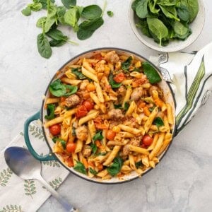 Sausage pasta in cast iron pan placed on benchtop, fresh spinach in a bowl and leaves scattered on the table.
