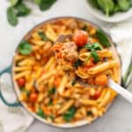 A big spoon holding up a scoop of sausage pasta with cast iron pan blurred in background with remainder of pasta.