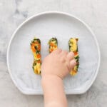 Three spinach omelette fingers on a child's silicone plate with a toddlers hand taking one of the fingers.