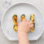 Three spinach omelette fingers on a child's silicone plate with a toddlers hand taking one of the fingers.