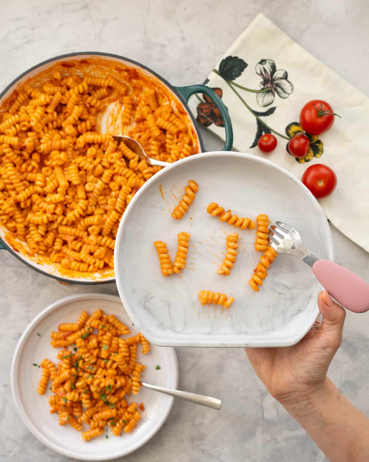 Woman's hand holding grey silicone baby plate with pieces of tomato pasta and baby fork with pink handle. Pasta in pan and on plate below in background.