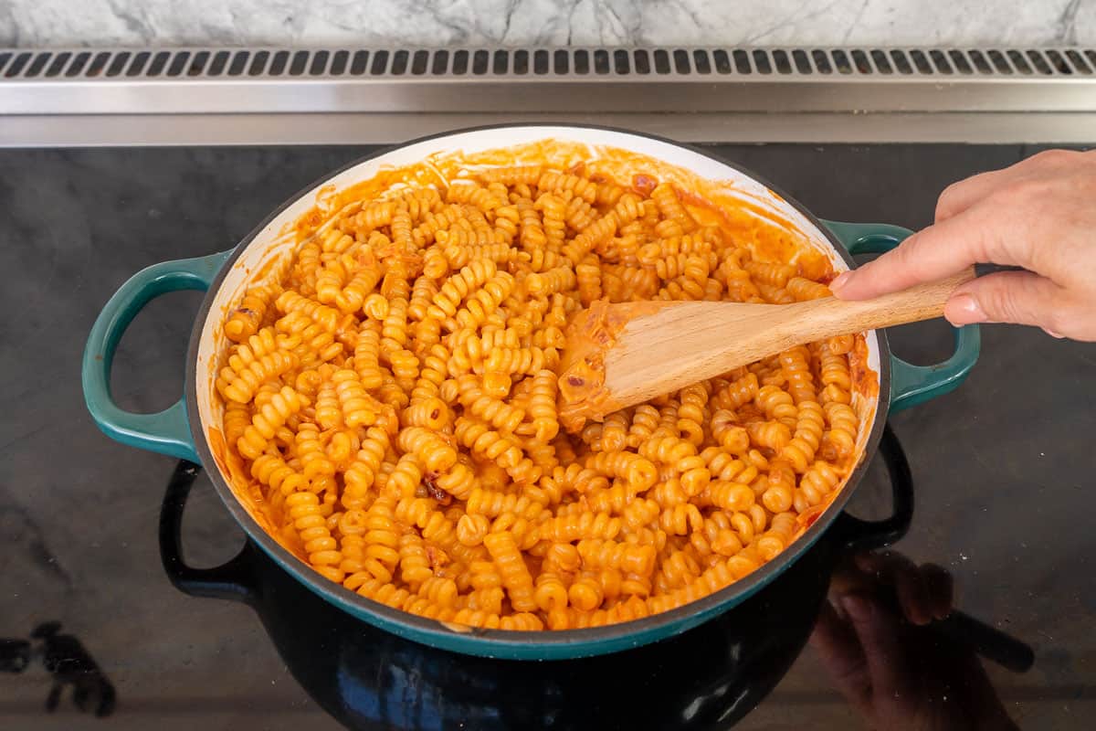 Hand holding wooden spoon mixing Creamy tomato pasta in skillet on stove top.