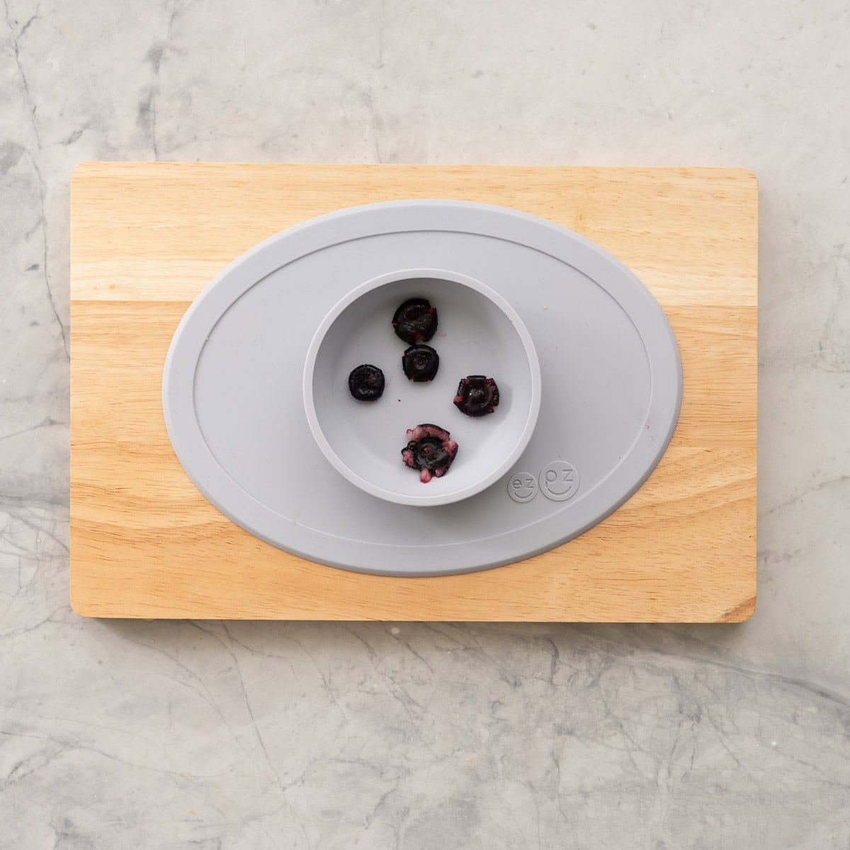 Silicone bowl on wooden chopping board with five blueberries inside, soft blueberries pushed down to be squashed.