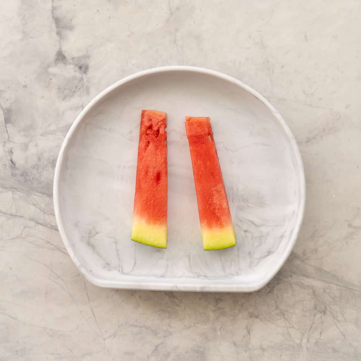 Two long rectangular slices of watermelon with skin at the bottom sitting on baby silicone plate.
