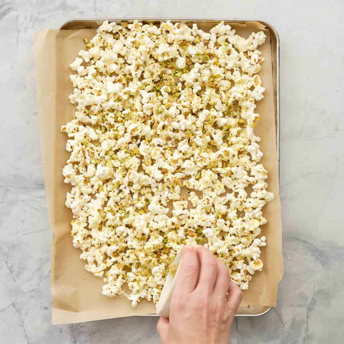 Hand sprinkling a small ceramic bowl of pistachios on top of tray of popcorn with melted white chocolate.