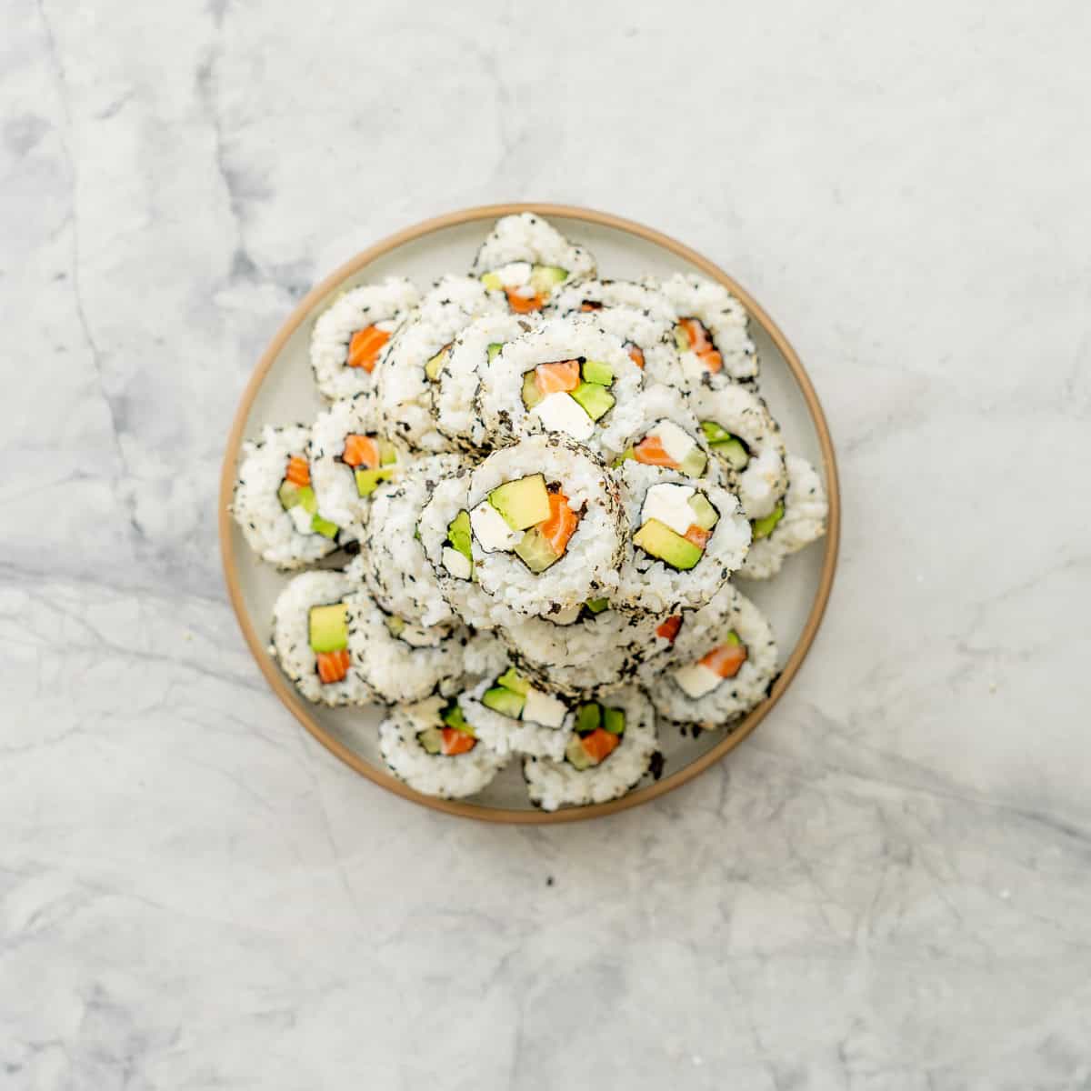 Snack of sushi in a tower face up showing fillings inside, cream cheese avocado, cucumber and salmon on a plate on benchtop.