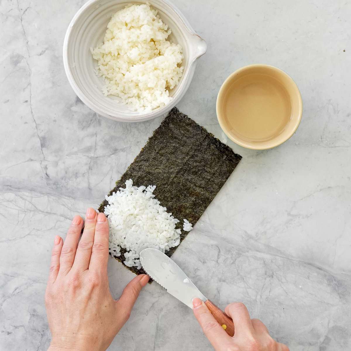 Sushi rice being pressed onto a sheet of nori with a spatula.