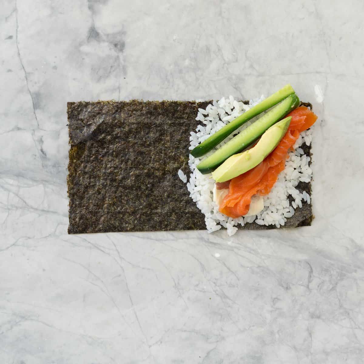 Sushi rice, salmon, avocado and sliced cucumber on half of a sheet of rectangular nori.