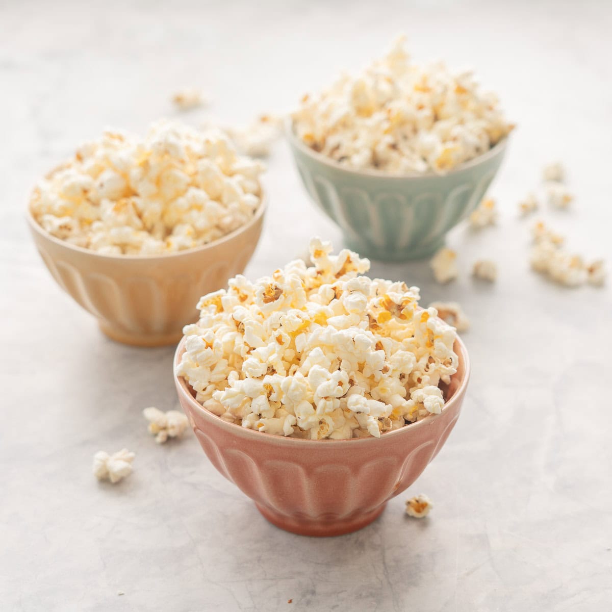 Three pastel coloured ceramic bowls filled with popcorn, sitting on a bench top with a few stray popcorn pieces.