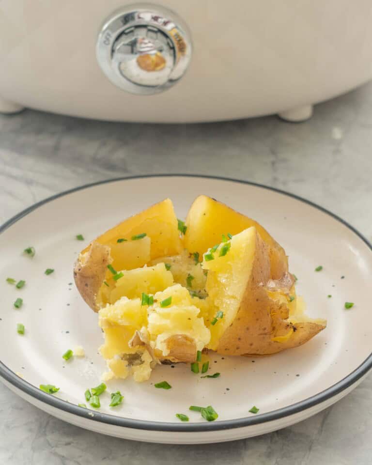 Slow cooker baked potato on a dinner plate cut in half with melted butter inside and sprinkled with chives.