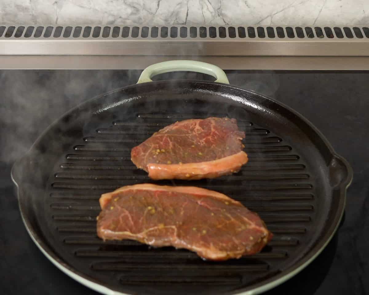 Two pieces of marinated steak on a ribbed cast iron pan on stove top.