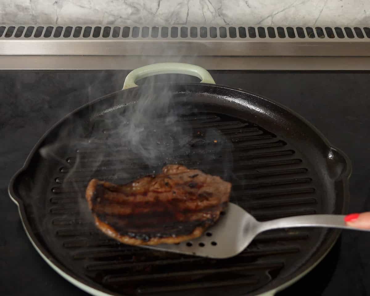 A piece of marinated steak being taken off a ribbed cast iron pan with metal spatula on stove top.