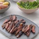 Marinated steak on a big plate sliced into thin pieces showing a medium cook inside, asparagus and broccolini in a bowl and seasoned potatoes on a bowl on bench top.