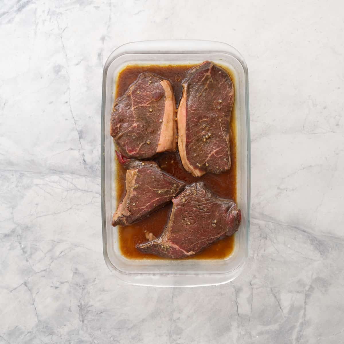 Four pieces of steak marinating in Steak marinade a glass dish on bench top.
