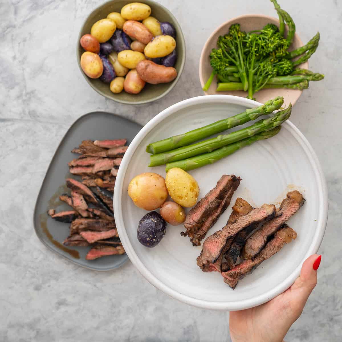 Dinner plate with marinated steak, potatoes and asparagus held up to camera. A big plate of marinated steak sliced into thin pieces showing a medium cook inside, bowl of broccolini and asparagus in a bowl and seasons potatoes in bowl on benchtop.