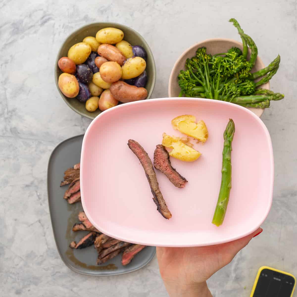 Baby silicone plate with two slices of marinated steak a halved potato and piece of asparagus held up to camera. A big plate of marinated steak sliced into thin pieces showing a medium cook inside, bowl of broccolini and asparagus in a bowl and seasons potatoes in bowl on benchtop.