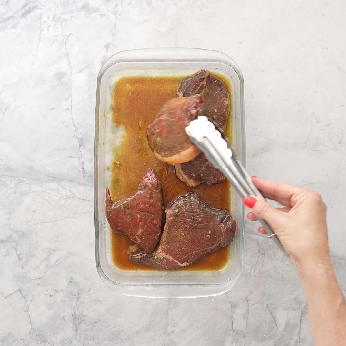 Four pieces of steak marinating in Steak marinade a glass dish on bench top. Hand holding tongs lifting and turning over a piece of steak.