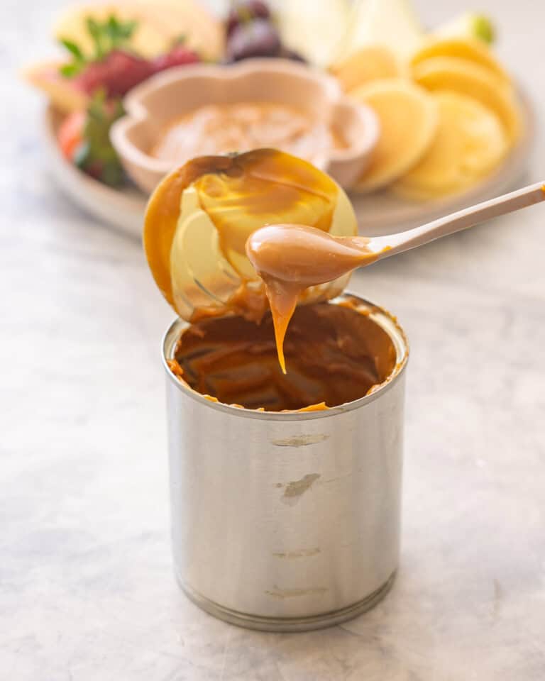 Spoonful of Condensed milk caramel being scooped out of can with lid taken off. Plate of Condensed milk caramel in small ramekin on plate with sliced apples, pears, grapes, strawberries and mini pancakes blurred in background.