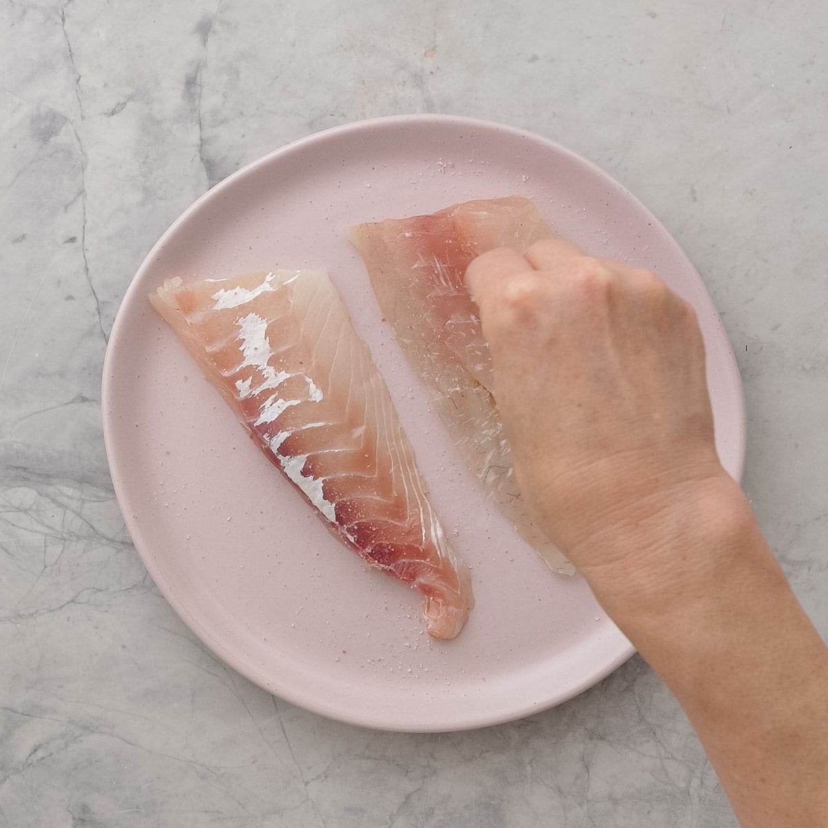 Hand sprinkling salt on top of two pieces of fish on a blush pink plate.