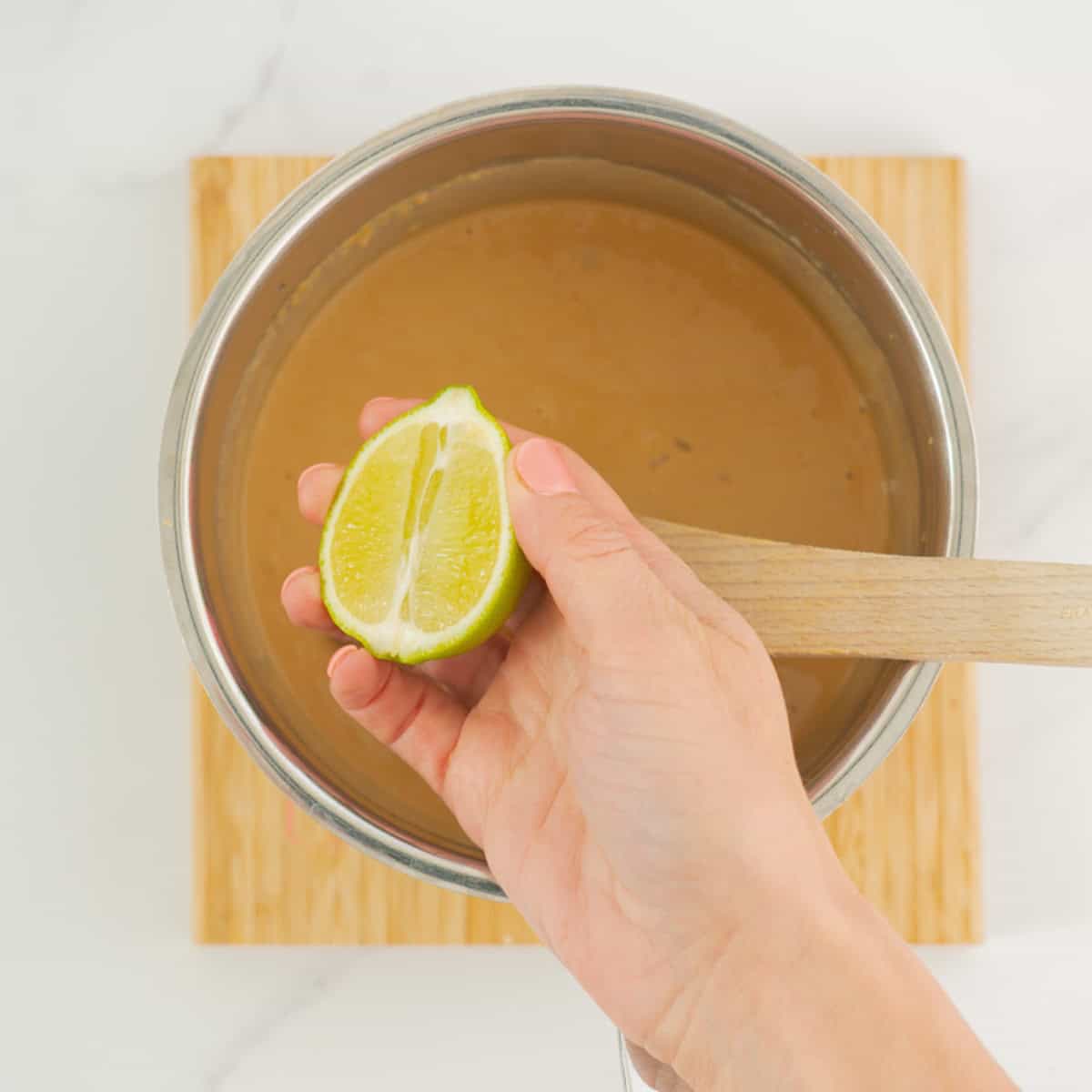 half a lime being held up above a sauce pan of smooth peanut sauce.