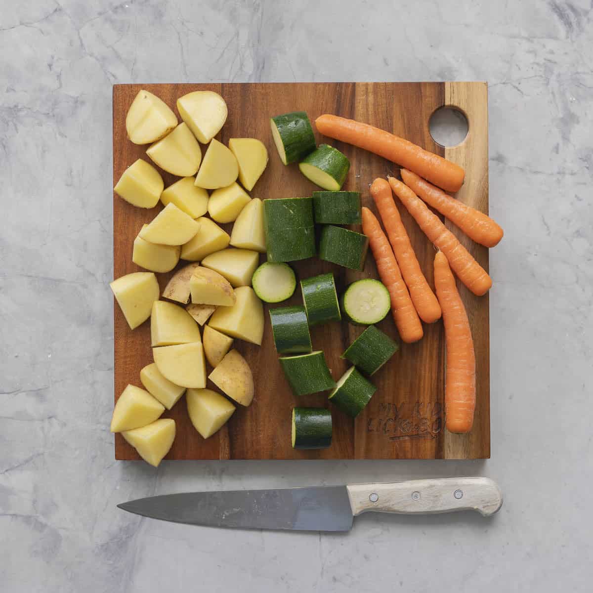 Wooden chopping board laid with chopped potatoes and zucchini in cubes and whole baby carrots. Knife on benchtop next to board.