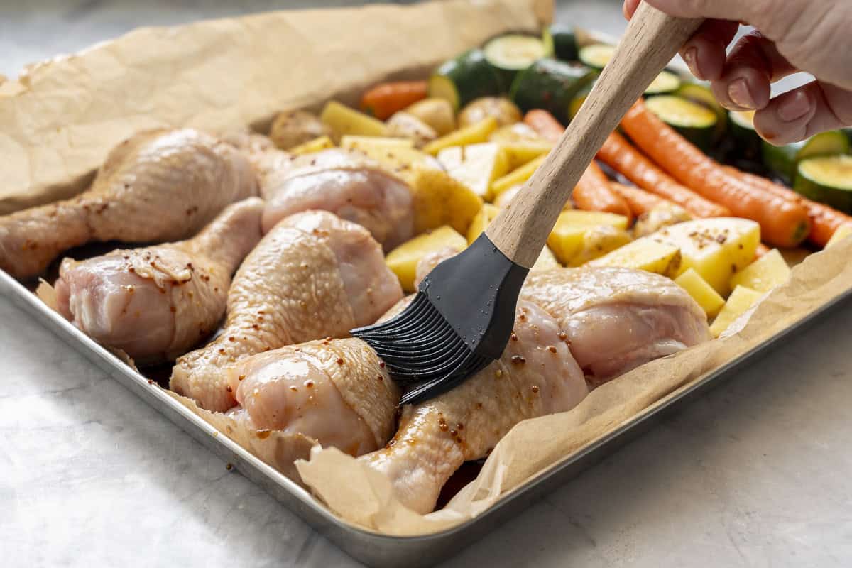Hand holding a basting brush with Honey soy sauce being brushed over chicken and vegetables on a baking tray lined with baking paper.