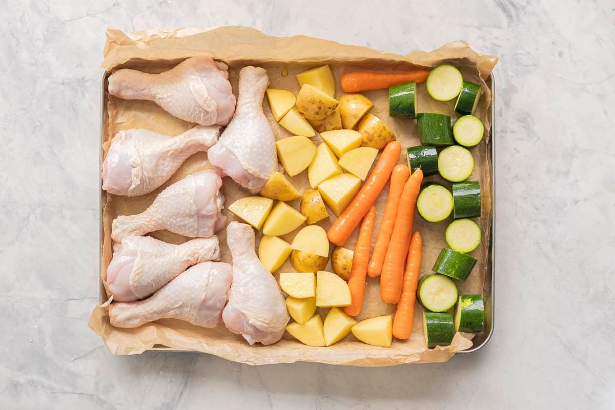 Chopped potatoes, zucchinis, whole carrots and six chicken drumsticks on a baking tray lined with baking paper.