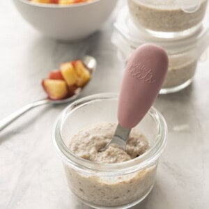 Apple chia pudding in small glass round container with pink handled toddler spoon inside, tablespoon resting on benchtop with chopped cooked apple and two small glass containers with chia pudding in background.