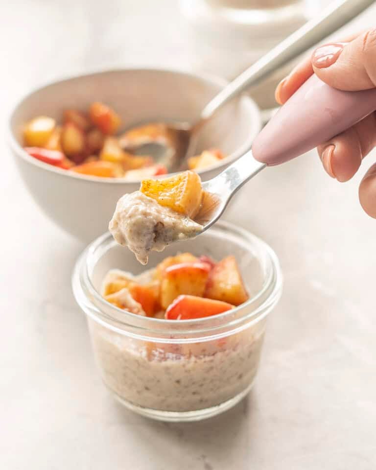 Small glass bowl on bench top with Apple Chia pudding and cooked spiced apple inside. Toddler spoon scooping pudding out of bowl.