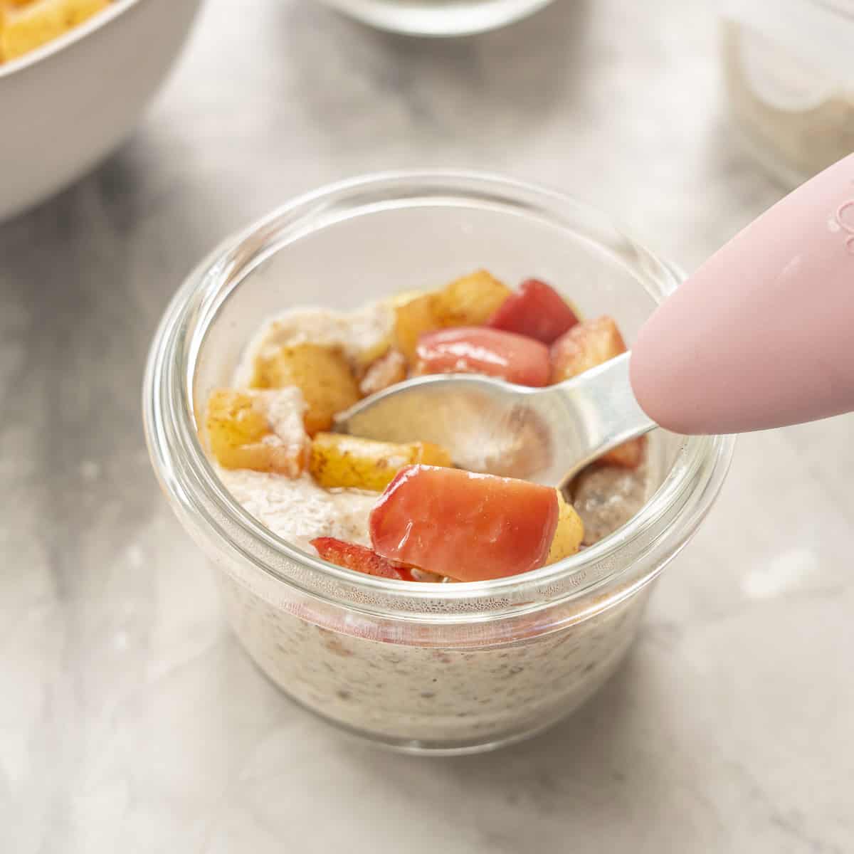Small glass bowl on bench top with Apple Chia pudding and cooked spiced apple inside. Toddler spoon scooping pudding out of bowl.