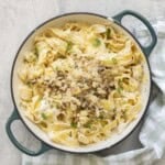 Cauliflower alfredo sauce mixed through cooked pasta topped with crispy roasted cauliflower, panko bread crumbs, thyme, parsley and parmesan cheese in a ceramic dutch oven pan on benchtop with green and white checkered tea towel.