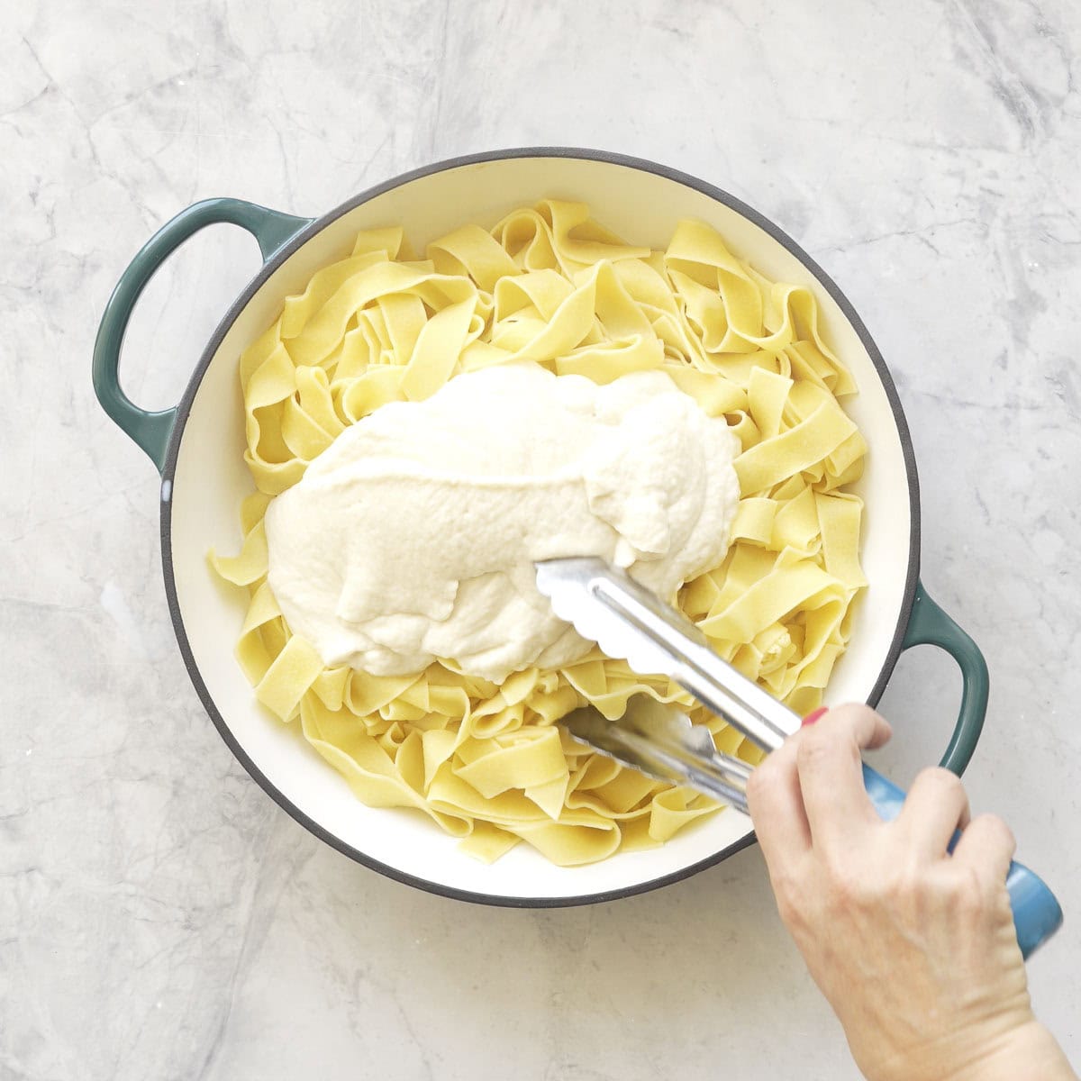 Cauliflower alfredo sauce poured on top of cooked pasta in ceramic dutch oven, hand holding tongs to mix.