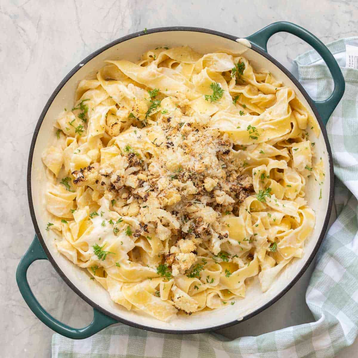 Cauliflower alfredo sauce mixed through cooked pasta topped with crispy roasted cauliflower, panko bread crumbs, thyme, parsley and parmesan cheese in a ceramic dutch oven pan on benchtop with green and white checkered tea towel.