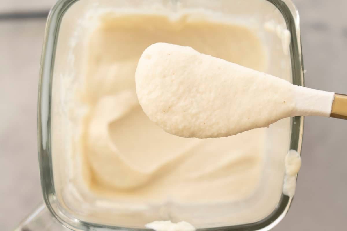 Spatula held up with thick creamy cauliflower alfredo sauce on it, blender with sauce in background below.