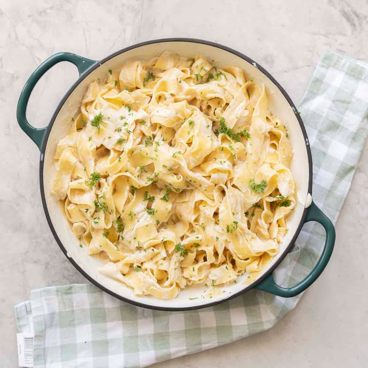 Cauliflower alfredo sauce mixed through cooked pasta topped with chopped parsley in a ceramic dutch oven pan on benchtop.