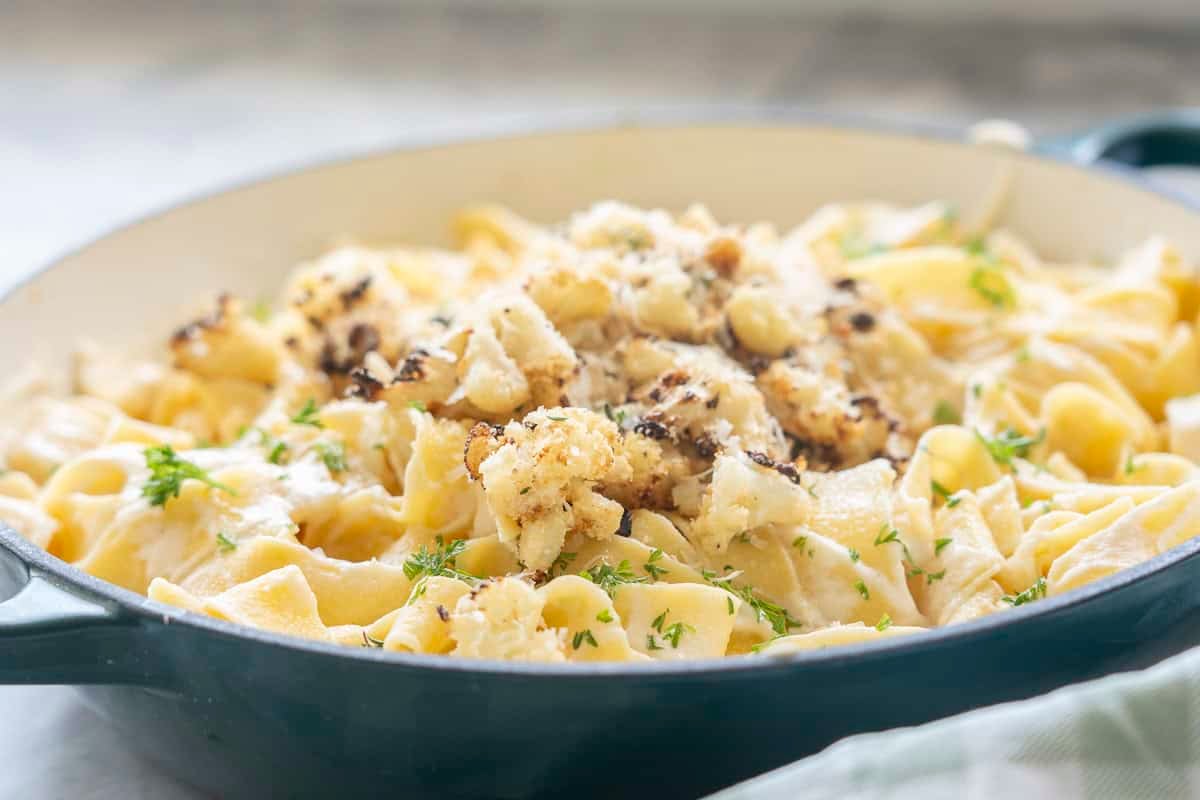 Cauliflower alfredo sauce mixed through cooked pasta topped with crispy roasted cauliflower, panko bread crumbs, thyme, parsley and parmesan cheese in a ceramic dutch oven pan on benchtop with green and white checkered tea towel.