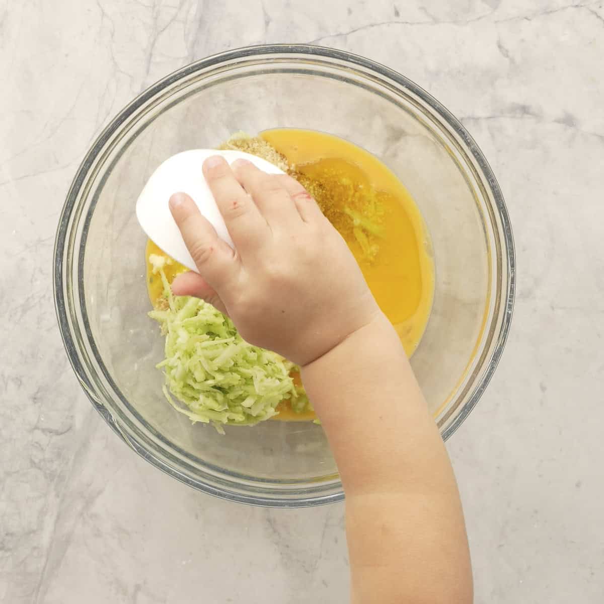 Toddler holding a small ramekin pouring ingredients into a bowl with other ingredients for chicken meatballs.
