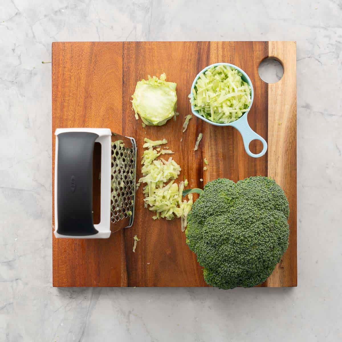 Whole broccoli head, grater, measuring cup of grated broccoli stalk on wooden chopping board.