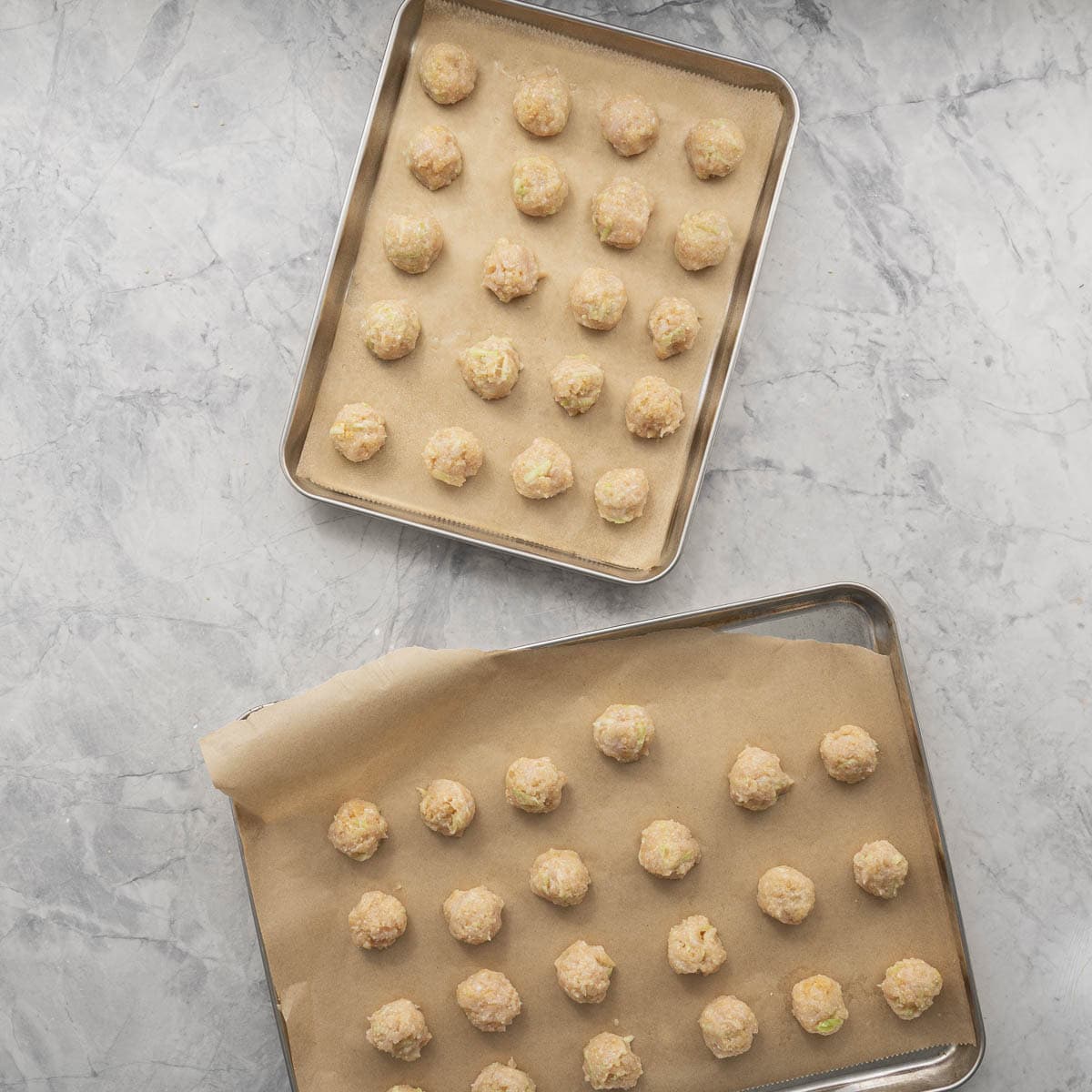 Two lined baking trays on benchtop with rolled uncooked chicken meatballs.
