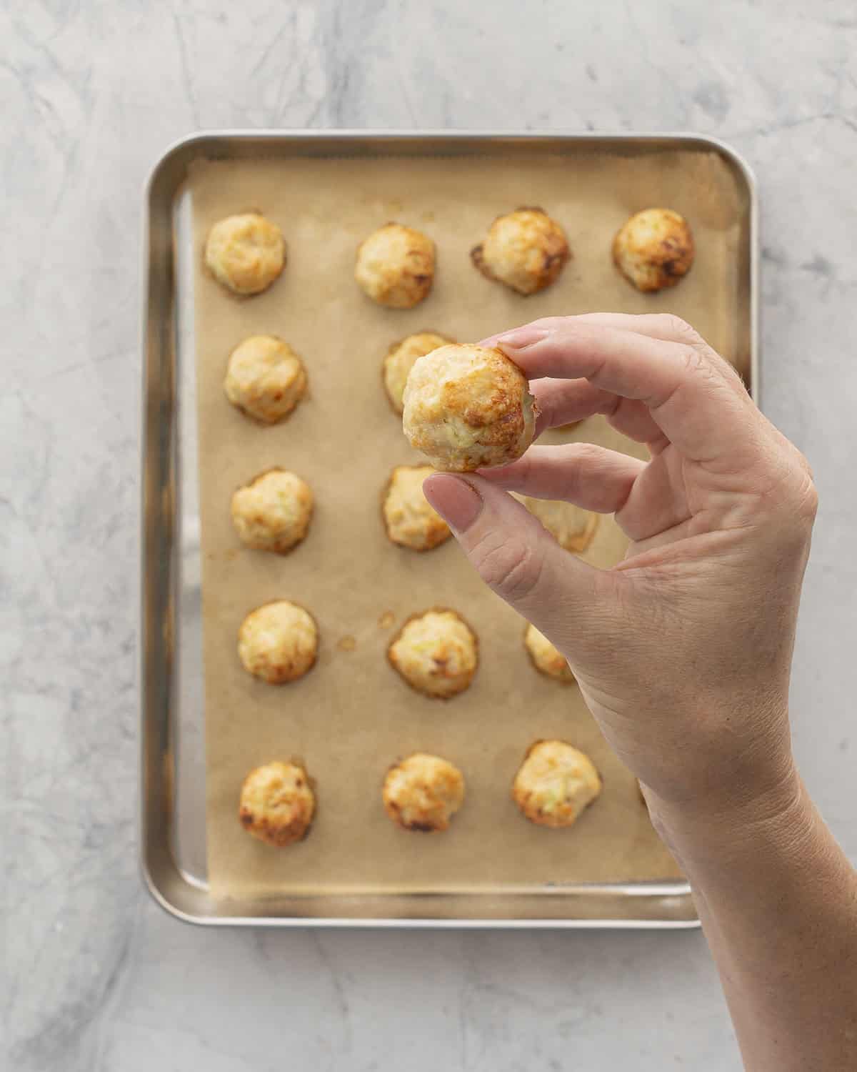 Hand holding up a chicken meatball above a lined baking tray of others on bench top.