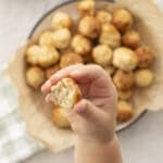 Toddlers hand holding up a chicken meatball bowl in background with baking paper with chicken meatballs inside, green checkered tea towel on benchtop. Sweet chilli sauce in a ramekin.