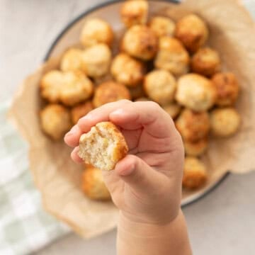 Toddlers hand holding up a chicken meatball bowl in background with baking paper with chicken meatballs inside, green checkered tea towel on benchtop. Sweet chilli sauce in a ramekin.