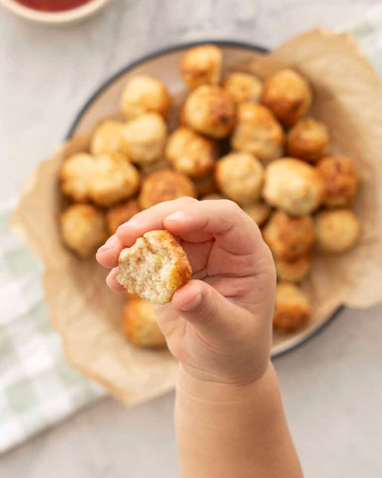 Toddlers hand holding up a chicken meatball bowl in background with baking paper with chicken meatballs inside, green checkered tea towel on benchtop. Sweet chilli sauce in a ramekin.