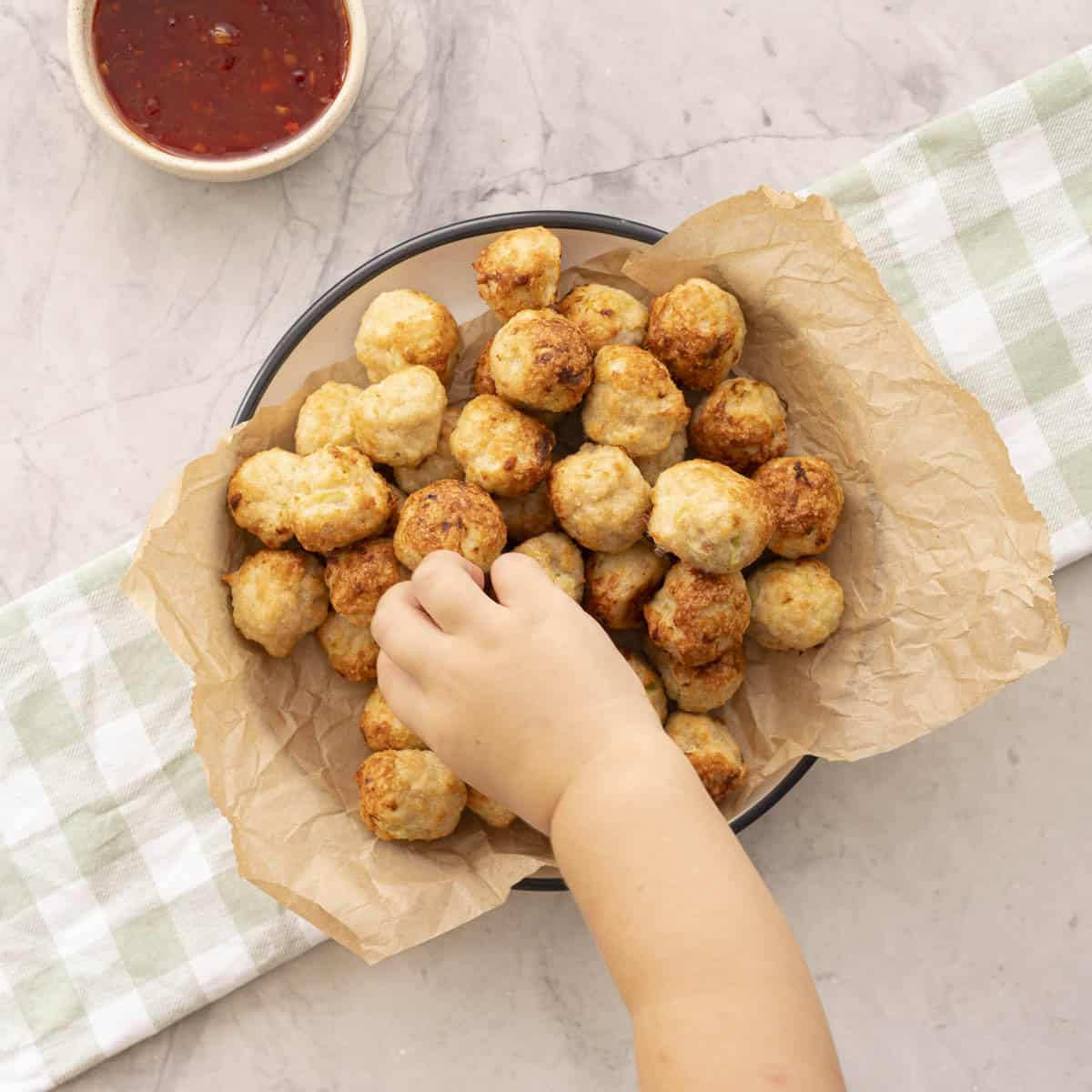 Toddlers hand reaching into a bowl lined with baking paper with chicken meatballs inside, green checkered tea towel on benchtop. Sweet chilli sauce in a ramekin.