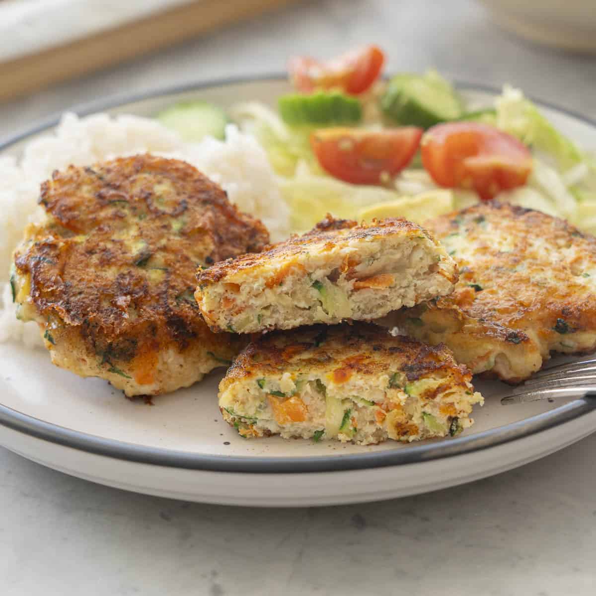 Three chicken patties, one cut in half showing the inside, a fresh salad and small portion of rice on a dinner plate.