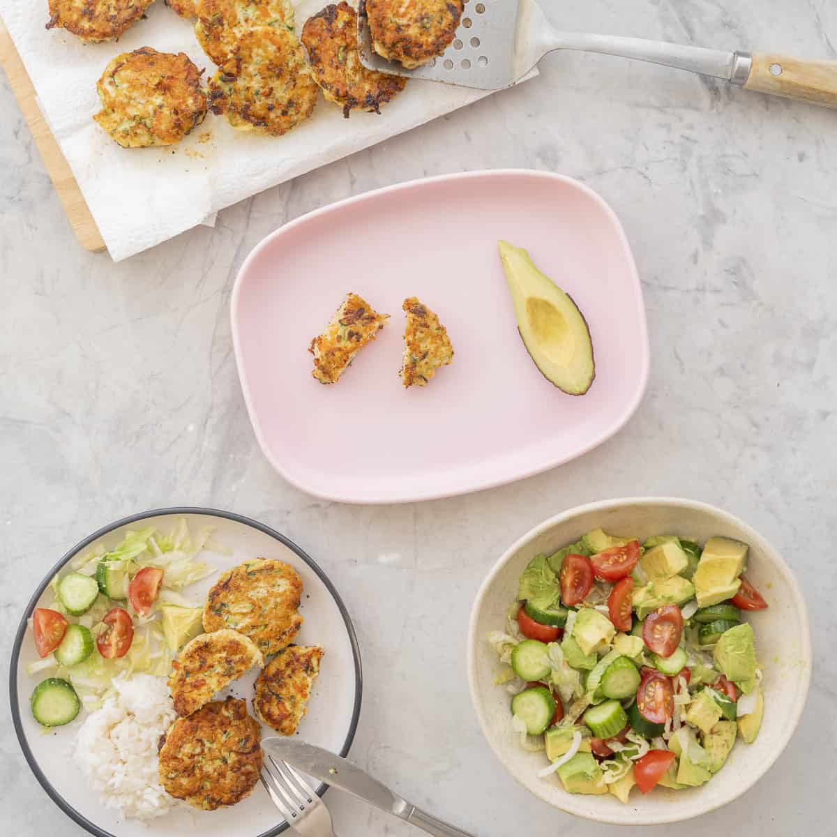 Three chicken patties, a fresh salad and small portion of rice on a dinner plate, silocone toddler plate with patty cut into two small pieces and a quarter avocado with half the skin removed, Bowl of salad and tray of chicken patties on benchtop.